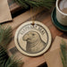 A decorative ornament featuring an elephant seal is displayed on a wooden surface, surrounded by pine branches and a cup of coffee.