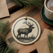 a decorative ornament with a deer illustration, placed on a wooden surface alongside a cup of coffee and some pine branches.