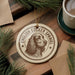 A circular ornament featuring an illustration of an English cocker spaniel dog, with the text "ENGLISH COCKER SPANIEL" around the edge. The ornament is placed on a wooden surface, with pine branches and a cup of coffee nearby.
