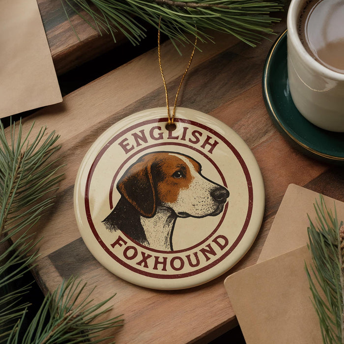 A circular ornament featuring a brown and white foxhound dog, with the text "ENGLISH FOXHOUND" printed on it, is displayed on a wooden surface alongside a cup of coffee and some pine branches.