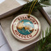 A decorative ornament in a box, featuring a circular design with a castle and waves, surrounded by greenery.