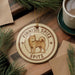A circular ornament featuring a brown dog, likely a Finnish Spitz, is displayed on a wooden surface alongside a cup of coffee and some pine branches.