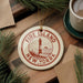 A circular ornament with the text "FIRE ISLAND NEW YORK" and a lighthouse image, placed on a wooden surface next to a cup of coffee and pine branches.