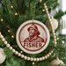 A Christmas tree decorated with ornaments, including a circular ornament with a portrait of a man wearing a hat and the word "FISHER" below it.