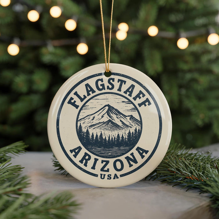 A circular ornament with the text "FLAGSTAFF ARIZONA USA" and a mountain illustration, hanging from a string against a blurred background of Christmas lights.