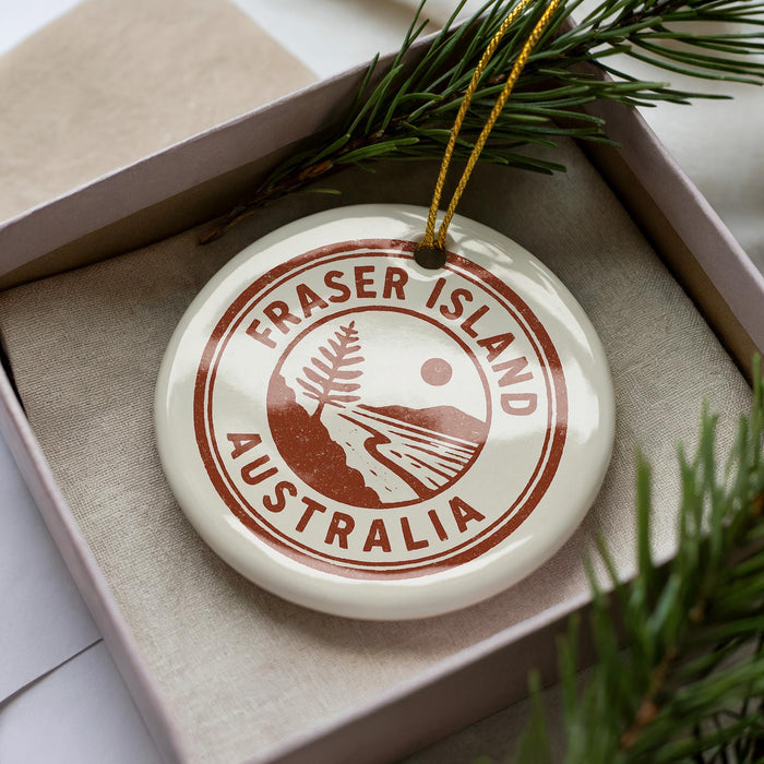 A white ornament with the text "FRASER ISLAND AUSTRALIA" printed on it, placed inside a box with pine branches.