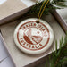 A white ornament with the text "FRASER ISLAND AUSTRALIA" printed on it, placed inside a box with pine branches.