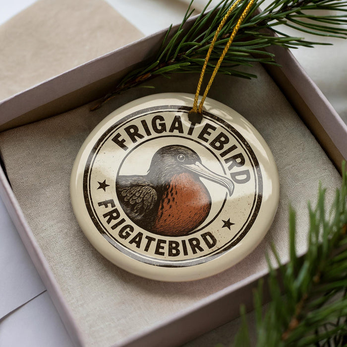 A circular ornament with the text "FRIGATEBIRD" and an illustration of a bird is displayed in a box, surrounded by pine branches.