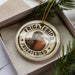 A circular ornament with the text "FRIGATEBIRD" and an illustration of a bird is displayed in a box, surrounded by pine branches.