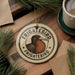 A circular ornament with the text "FRIGATEBIRD FRIGATEBIRD" and an illustration of a bird is displayed on a wooden surface, surrounded by pine branches and a cup of coffee.