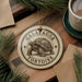 A round ornament featuring a tortoise, with the text "GALAPAGOS TORTOISE" printed on it, is displayed on a wooden surface alongside a cup of coffee and some pine branches.