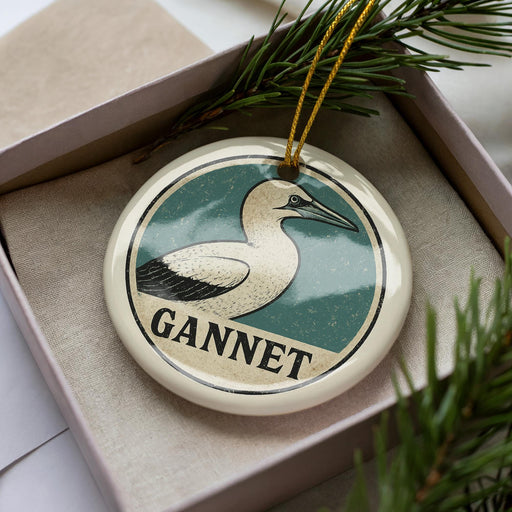A circular ornament with a bird image and the word "GANNET" printed on it, placed in a box with pine branches.
