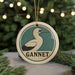 A circular ornament with a bird image and the word "GANNET" written on it, hanging in front of a Christmas tree with twinkling lights.