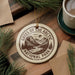 A circular ornament with the text "GATES OF THE ARCTIC NATIONAL PARK" and a mountain landscape design, placed on a wooden surface next to a cup of coffee and pine branches.