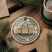 A circular ornament with the words "GATEWAY ARCH NATIONAL PAARK" printed on it, featuring an illustration of the Gateway Arch in St. Louis, Missouri. The ornament is hanging from a string, and there is a cup of coffee and some pine branches in the background.