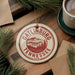 A circular ornament with the text "GATLINBURG TENNESSEE USA" printed on it, surrounded by pine branches and a cup of coffee on a wooden surface.