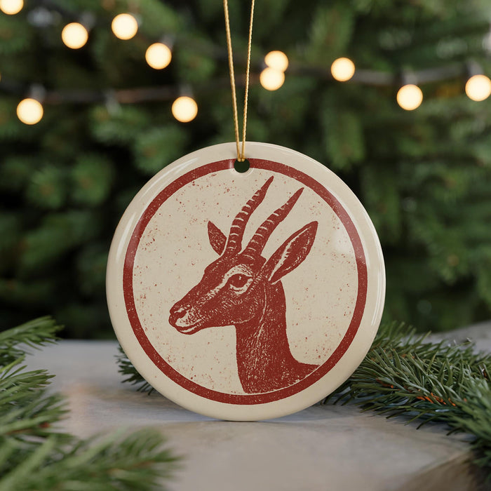A round ornament featuring a red and white deer head design is displayed in front of a blurred Christmas tree with twinkling lights.