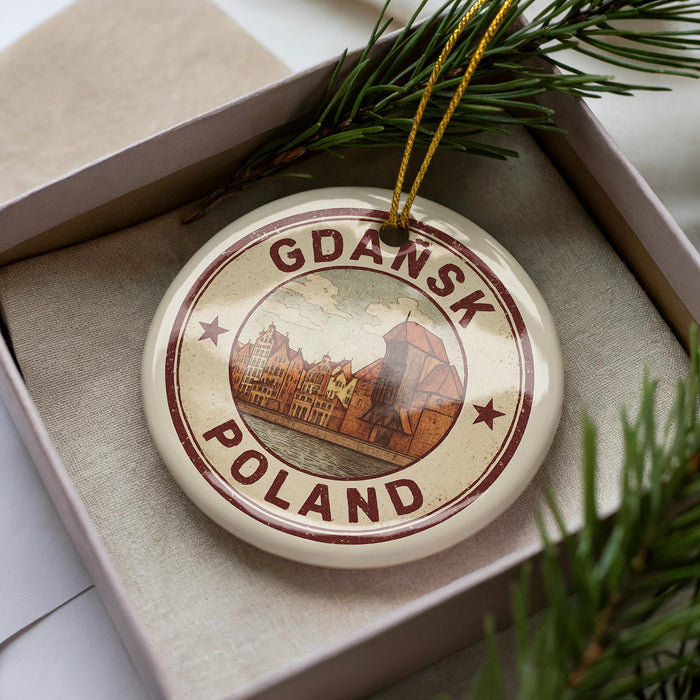 A circular ornament with the text "Gdansk Poland" printed on it, placed inside a box with pine branches.
