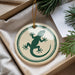 A round ornament with a green lizard design is displayed in a box, surrounded by pine branches.