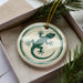 A ceramic ornament in the shape of a gecko, with a green and white design, is displayed in a box with pine branches.