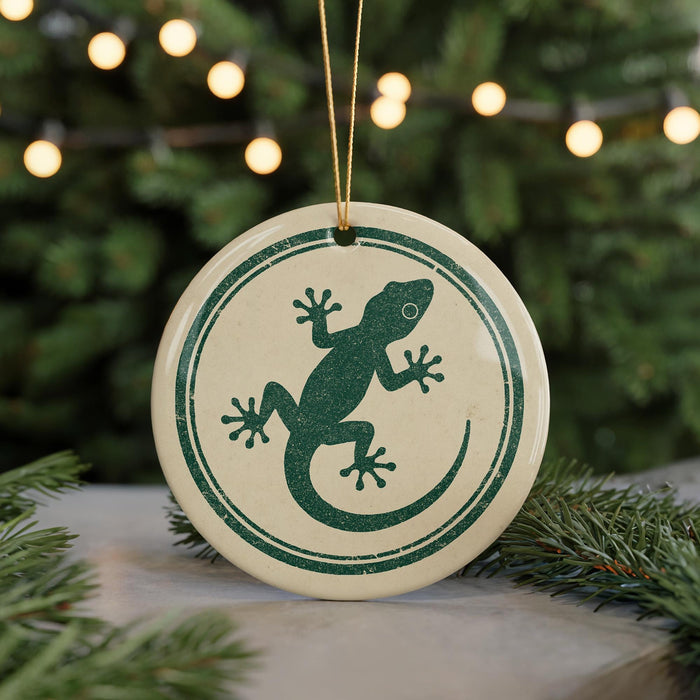 A round ornament with a green lizard design is displayed against a blurred background of a Christmas tree with lights.