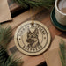 A round ornament featuring a German Shepherd dog's head, with the text "German Shepherd" around the edge, is displayed on a wooden surface alongside a cup of coffee and some pine branches.