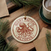 A round ornament featuring an illustration of an octopus is displayed on a wooden surface, surrounded by pine branches and a cup of coffee.
