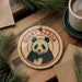 A panda ornament with the text "GIANT PANDA" is displayed on a wooden surface, surrounded by pine branches and a cup of coffee.