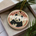 A circular ornament with a panda bear image and the text "GIANT PANDA" printed on it, placed inside a box with pine branches.