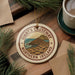 A circular ornament with the text "Giant's Causeway Northern Ireland" on it, surrounded by pine branches and a cup of coffee on a wooden surface.