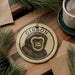 A ceramic ornament featuring a monkey's face is displayed on a wooden surface, surrounded by pine branches and a cup of coffee.