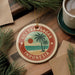 A circular ornament with a palm tree and beach scene, surrounded by pine branches, sits on a wooden surface.