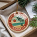 A round ornament with a tropical beach scene and palm trees, featuring the text "Gili Trawangan Indonesia" on it, is displayed in a box with pine branches.