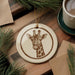 A ceramic ornament featuring a giraffe's head is displayed on a wooden surface, surrounded by pine branches and a cup of coffee.
