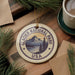 A circular ornament with the text "GLACIER NATIONAL PARK USA" and a mountain landscape design hangs on a wooden surface, surrounded by pine branches and a cup of coffee.
