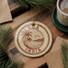 A circular ornament with the text "Goa India" is displayed on a wooden surface, surrounded by pine branches and a cup of coffee.