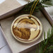 A round ornament featuring a close-up of a golden eagle's head, displayed in a box with pine branches.