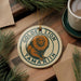 A round ornament featuring a golden lion, a type of primate, is displayed on a wooden surface alongside a cup of coffee and some pine branches.