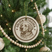 A round ornament with a golden retriever dog's face is hanging on a Christmas tree, surrounded by other ornaments and lights.