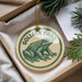 A circular ornament featuring a green frog, with the text "GOLIATH FROG" printed on it, is displayed in a box with pine branches.