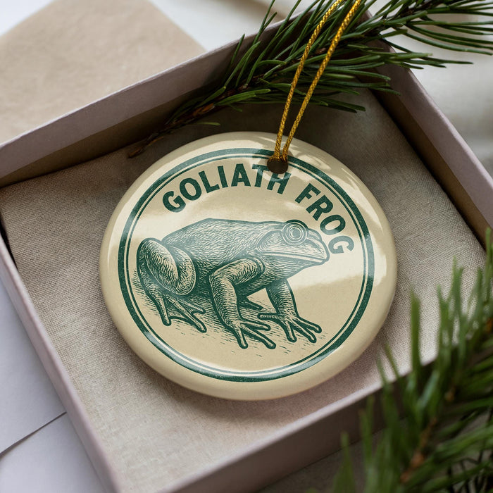 A circular ornament with a green frog image and the text "GOLIATH FROG" on it, placed in a box with pine branches.