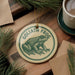 A circular ornament featuring a green frog on a wooden surface, with the text "GOLIATH FROG" printed on it.