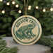A circular ornament featuring a detailed illustration of a frog, with the text "GOLIATH FROG" printed on it. The ornament is hanging from a string, and there are pine branches visible in the background.