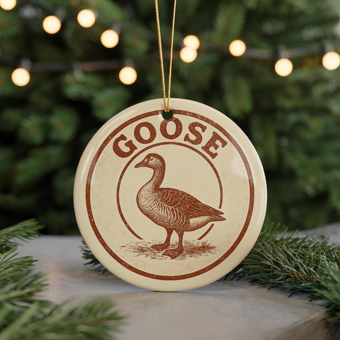 A circular ornament with the word "GOOSE" and a graphic of a goose is displayed on a table, surrounded by pine branches and twinkling lights.