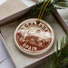 A circular ornament with the text "Granada, Spain" printed on it, placed inside a box with pine branches.