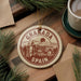 A circular ornament with the text "GRANADA SPAIN" is displayed on a wooden surface, surrounded by pine branches and a cup of coffee.