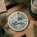 A circular ornament with the text "GRAND CAMERON Cayman Islands" and a palm tree design, hanging on a wooden surface.