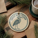 A circular ornament featuring a blue heron, with the text "GREAT BLUE HERON" printed on it, is displayed on a wooden surface alongside a cup of coffee and some pine branches.
