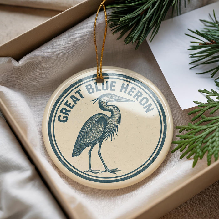 A circular ornament with a blue heron illustration and the text "GREAT BLUE HERON" is displayed in a box with pine branches.