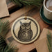 a round ornament with an owl design, placed on a wooden surface next to a cup of coffee and some pine branches.